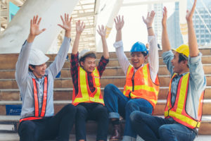 A group of Gen Z construction workers in safety vests and hard hats raising their hands in celebration while sitting on steps at a job site, representing hiring Gen Z in the trades.